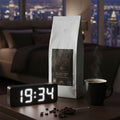 A bag of coffee on a desk top next to a digital clock