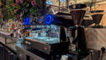 A professional espresso machine with blue LED ring lights and a matching coffee grinder on a stainless steel bar counter in a decorated cafe setting.